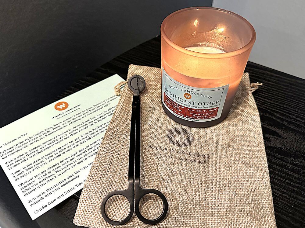 Aroma candle lit and staged on burlap bag with wick trimmers and candle care card.