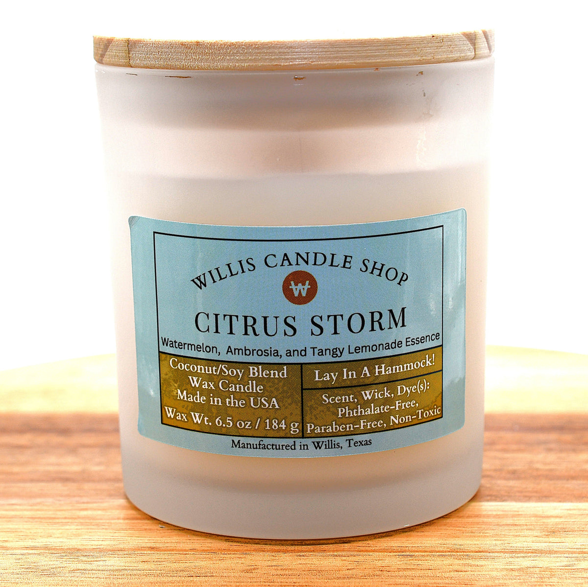 A frosted glass jar candle with a light wood lid sits on a wooden surface against a white background. The front label reads 'WILLIS CANDLE SHOP CITRUS STORM' with a light blue background and brown text blocks.