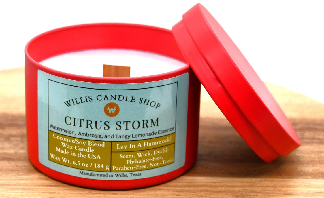 A red wax candle in a red metal tin sits on a wooden surface with its lid leaning against the side. The front label reads Willis Candle Shop and Citrus Storm.